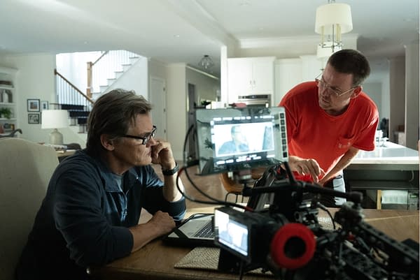 A scene depicting two men discussing a film project in a home setting. One man is seated at a table with a laptop, looking thoughtful, while the other is standing next to a camera setup, pointing to the equipment.