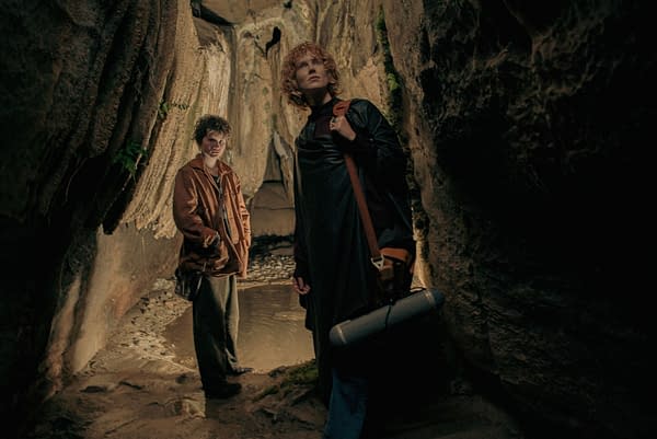 Two people are exploring a narrow cave; one is a child with short hair in a brown jacket, while the other is an adult with curly hair wearing a black coat, holding a bag and a portable speaker. The rocky walls and a small pool of water create a mysterious atmosphere.