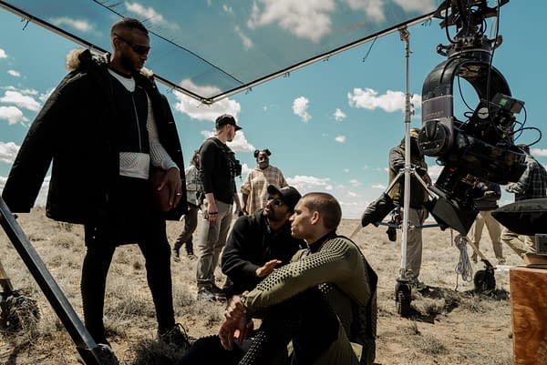 On the set of the film 'HIM', actors Marlon Wayans and Tyriq Withers are seen with crew members in a desert landscape, preparing for a scene. A large camera setup is visible, capturing the behind-the-scenes action.