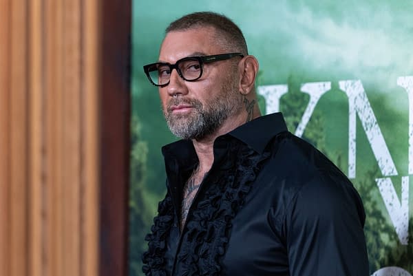 An actor with a beard and glasses, dressed in a black shirt with ruffles, poses at a movie premiere event for 'Knock at the Cabin', set in front of a green background featuring the film's title.