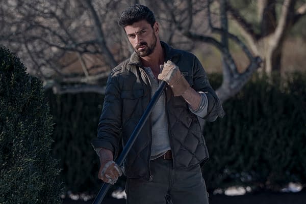 Michele Morrone as Enzo in The Housemaid, standing outdoors with a focused expression while holding a long tool, dressed in a quilted jacket and gloves, with a blurred background of trees and shrubs.