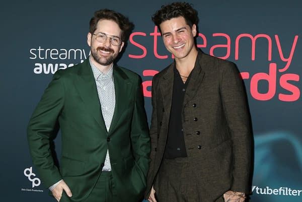 Two male celebrities pose for a photo at the Streamy Awards, with one wearing a green suit and the other in a dark, stylish outfit. They are standing against a backdrop featuring the event's logo.