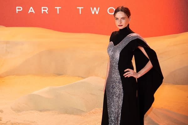 A woman wearing a glamorous black gown with a sparkling silver accent poses confidently on the red carpet at a film premiere, set against a backdrop of sandy dunes and the words "PART TWO" in the background.