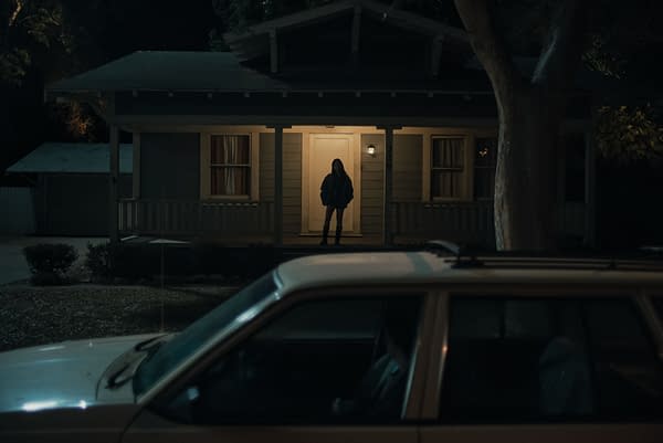 A woman stands in front of a lit house at night, wearing a dark jacket and jeans, while a car is parked nearby in the foreground. The scene conveys a sense of intrigue and tension, reflecting themes from the movie 'Obsession.'