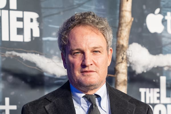 A close-up of a man with curly gray hair and a serious expression, dressed in a black suit and tie, standing in front of a backdrop related to 'A Quiet Place' film series. The background features snowy trees and promotional text.