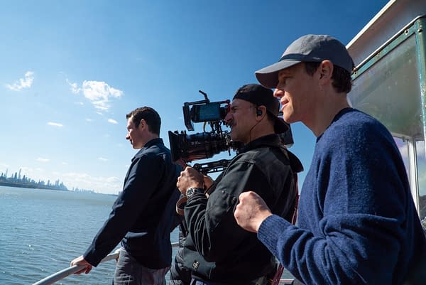Film crew on a boat filming a waterfront skyline with a large cinema camera. Three men stand along the railing under a bright blue sky.