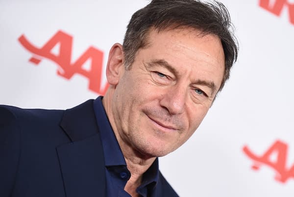 British actor Jason Isaacs smiles at a red carpet event, wearing a navy suit with a blue shirt, against a backdrop featuring a prominent logo.