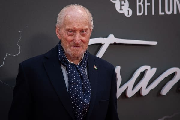 A man with a scruffy beard and light hair, wearing a polka dot scarf and a dark blazer, poses on a red carpet at an event in London. The background features the name of the film in stylized lettering, indicating a glamorous premiere atmosphere.