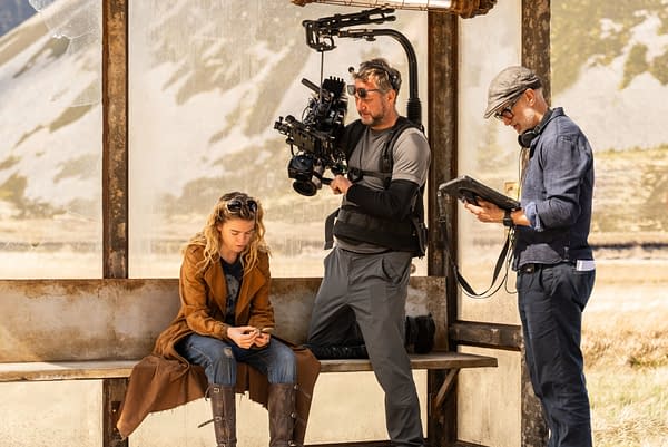 On the Supergirl set, a blonde actress in a brown coat sits on a bench while a camera operator films her and a crew member reviews a tablet nearby.