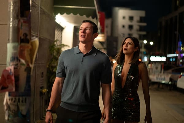 A man in a blue polo and a woman in a sparkly black dress walk through a brightly lit city street at night, both looking upward in surprise.