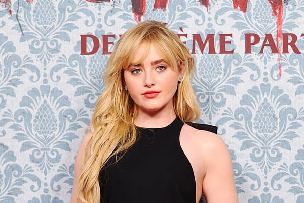 A woman with long, wavy blonde hair stands in front of a blue damask-patterned backdrop, wearing a black dress. She has a neutral expression and is accessorized with stud earrings.