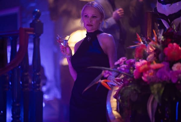 Pamela Anderson portrays Beth Davenport in a scene from 'The Naked Gun', wearing a black dress and holding a cocktail, set against a background of vibrant lighting and floral arrangements.