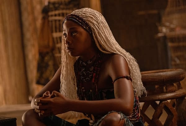 Thuso Mbedu as Zélie sits in a warm, rustic setting in Children of Blood and Bone, wearing long light braids and beaded accessories.
