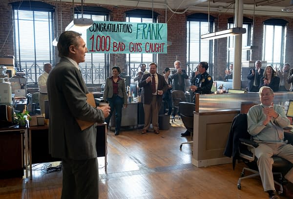 In a celebratory office scene from The Naked Gun, Liam Neeson as Frank Drebin Jr. stands in front of a banner congratulating him for catching 1,000 bad guys, while coworkers applaud and smile around him.