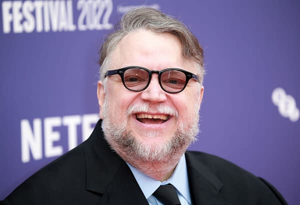 A portrait of Guillermo del Toro, the acclaimed Mexican filmmaker, smiling widely while wearing black glasses and a dark suit. The background features a purple banner for a 2022 festival.