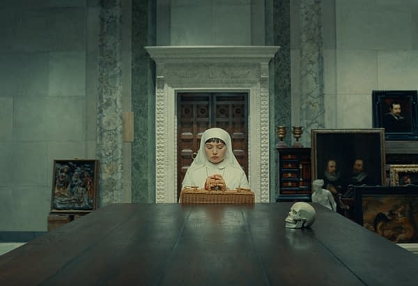 A scene from 'THE PHOENICIAN SCHEME' featuring Mia Threapleton as Liesl, dressed in a white nun's habit, sitting at a long wooden table with her hands clasped over a basket. The background showcases an ornate room decorated with various artworks, an unusual skull placed on the table, emphasizing a sense of contemplation.