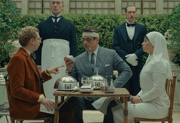In a scene from the film 'The Phoenician Scheme,' Michael Cera as Bjorn and Benicio Del Toro as Zsa-Zsa Korda engage in conversation at a dining table, flanked by two waitstaff. Del Toro's character is depicted bandaged with a cigarette in his mouth, while a nurse, portrayed by Mia Threapleton, sits to the right.