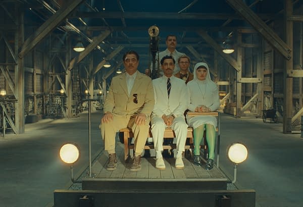 A scene from the film "The Phoenician Scheme," featuring actors seated together in a minimalist industrial setting. The characters include a man in a beige outfit, a man in a white suit, a woman in a white dress and headscarf, and others, all depicted with a stylized, cinematic aesthetic.