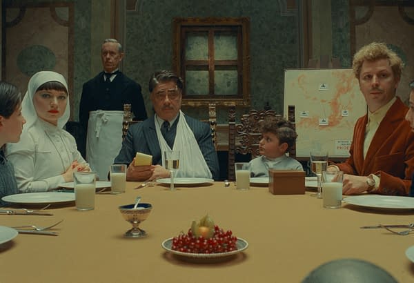 A scene from 'The Phoenician Scheme' featuring a formal dining table with six characters, including Mia Threapleton, Benicio Del Toro, and Michael Cera, in a vintage setting. The characters display a range of expressions, while a map and decorative dish are visible in the background.