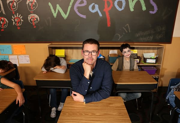 A man sits at a school desk with a thoughtful expression, surrounded by children in a classroom setting. The blackboard behind him features the word 'WEAPONS' in colorful letters, while some children bow their heads or look away, evoking a sense of somberness and reflection.