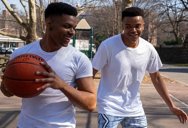 Two young men are playing basketball in an outdoor court, smiling and enjoying the game. One of them is holding a basketball while wearing a plain white t-shirt.