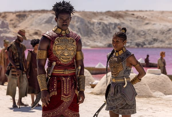 Damson Idris and Cynthia Erivo stand in ornate warrior costumes on a desert set for Children of Blood and Bone, with other figures and a pink lake in the background.