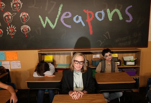 In a classroom setting, a woman with glasses sits at a desk with a serious expression, while two students exhibit contrasting behaviors: one slumped over, hidden beneath their arms, and another smiling with face paint. The chalkboard behind them displays the word 'Weapons' in colorful letters.