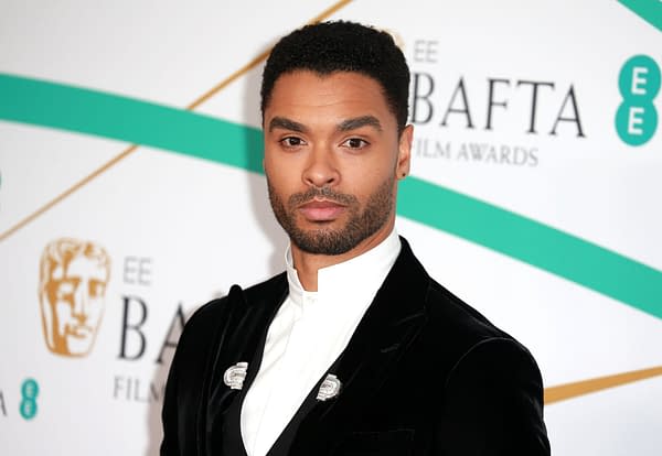 A portrait of Regé-Jean Page at a red carpet event, wearing a black velvet suit and a white shirt. He is looking directly at the camera against a backdrop featuring the BAFTA logo.