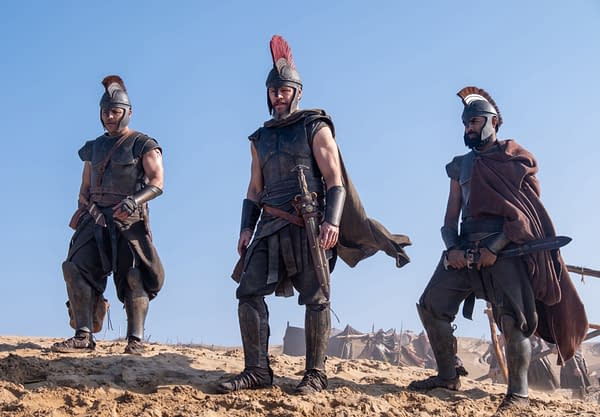 Matt Damon, Himesh Patel, and Jimmy Gonzales stand in armor as ancient Greek warriors in The Odyssey, with swords at their sides under a clear blue sky.