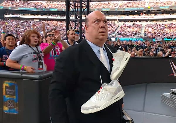 A suited man, Paul Herman, appears confused as he stands at a wrestling event, with a pair of white sneakers draped over his shoulder. The crowd in the background is lively, with fans cheering and waving their hands.