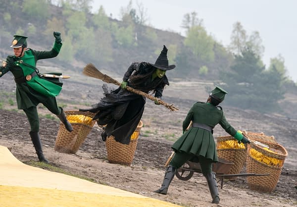 A scene from 'Wicked: For Good' featuring Cynthia Erivo as Elphaba, portrayed in a dramatic pose while flying on her broomstick, wearing a dark dress and a pointed hat. Two characters in green costumes appear to chase her, adding dynamic action to the moment.