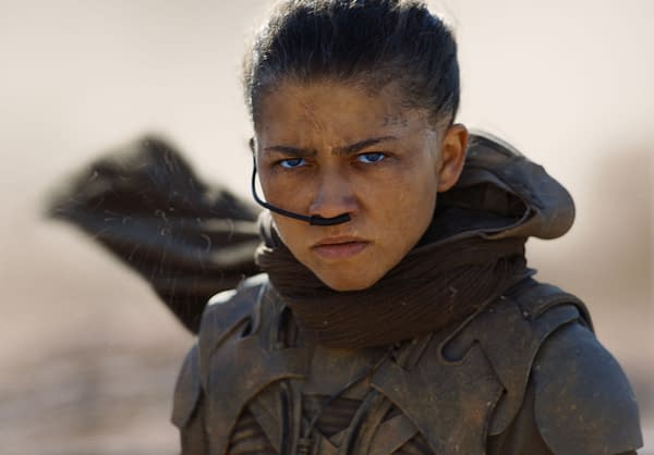 A determined young character from 'Dune' featuring intense blue eyes and a serious expression, dressed in a weathered outfit with a scarf and a futuristic facial device. The background suggests a harsh desert environment.