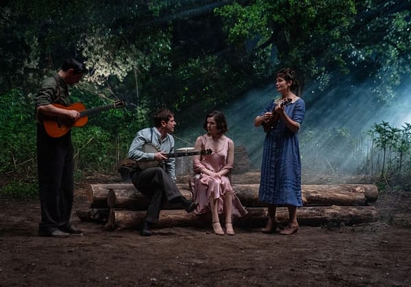 A scene featuring four performers in a forest setting, with two holding string instruments and two seated, creating a cozy atmosphere illuminated by soft, natural light filtering through the trees.