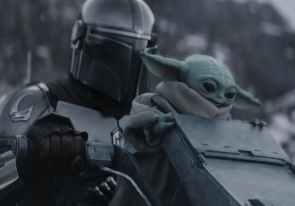 The Mandalorian, wearing a silver helmet and armor, holds onto a vehicle while Grogu, a small green creature with large ears, sits beside him wrapped in a grey robe. The scene conveys a moment of adventure in a snowy landscape.