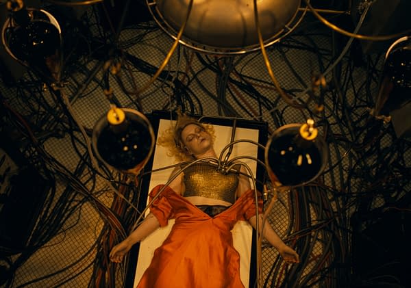A surreal scene featuring a woman with blonde hair lying on a table, wearing a golden top and a long red dress, surrounded by intricate wires and overhead lights, creating a dramatic and otherworldly atmosphere.