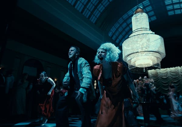 A dramatic scene featuring a man and a woman dancing energetically in a lavish party setting, illuminated by a large chandelier. The woman, wearing a red dress and with curly hair, appears to be passionately expressing herself, while the man beside her smiles intensely, creating a lively atmosphere.