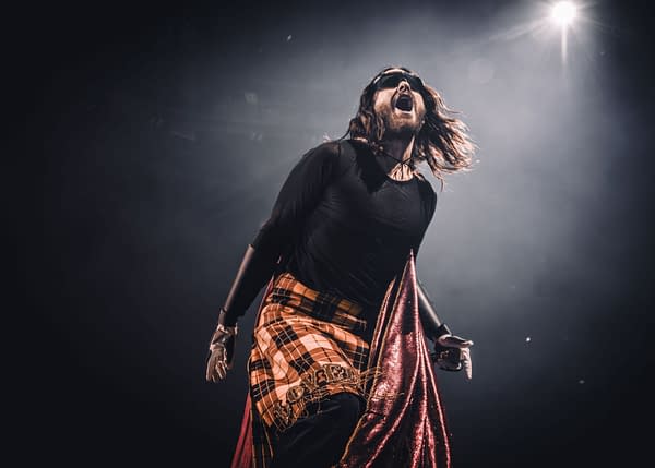 Jared Leto of Thirty Seconds to Mars performs energetically on stage, wearing a vibrant outfit with a plaid skirt and a flowing cape. Stage lights illuminate the scene, capturing the excitement of a live music concert.