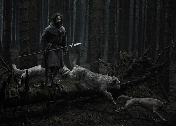 <strong>Aaron Taylor-Johnson</strong> stands in a dark forest holding a spear, with two wolves at his feet in a scene from <em>WERWULF</em>.