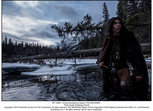 Forrest Goodluck as Hawk in THE REVENANT, standing in a partially frozen river with snowy terrain and dark trees in the background under a cloudy sky.