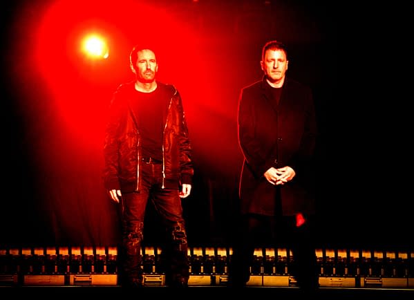 Trent Reznor and Atticus Ross stand on stage under a dramatic red light during the Disney Entertainment Showcase at D23, showcasing their fashion and presence in front of an audience.