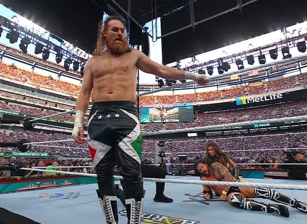 A wrestler with a prominent red beard and long hair, Sami Zayn, stands in the wrestling ring, wearing black pants adorned with a colorful design. The background features an excited crowd at a large stadium during WWE SummerSlam.