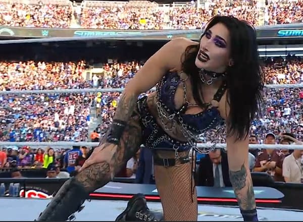 A wrestler with dark hair and striking makeup, Rhea RIpley, is kneeling in a ring, wearing a chains-adorned outfit in front of a large audience. The atmosphere conveys intensity and excitement at WWE SummerSlam, where Ripley just lost to Naomi.