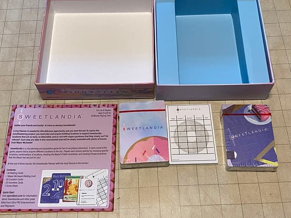 An array of the few components that make us UltraPro Entertainment and Stoneblade Entertainment's candy card game, Sweetlandia. Contents include a rules pamphlet, two decks of cards, and a pad to keep track of scores in-game.