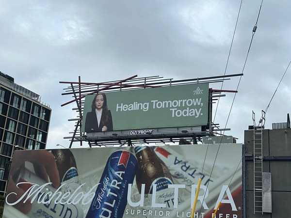 A billboard displays a professional woman with long dark hair in a black suit, promoting the message "Healing Tomorrow, Today." The billboard is set against a cloudy sky, with other advertisements for Michelob Ultra visible below.