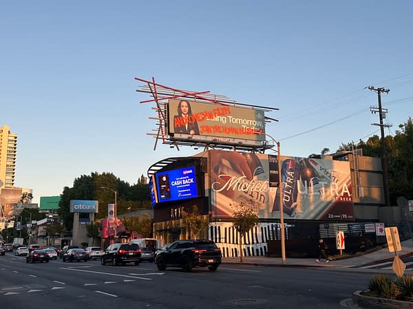 A vibrant street scene featuring a large billboard advertising a comedy show with the text 'Andromeda Jon' and a promotional message about cash back from a bank on another display. Various cars are seen driving along the road, with buildings and trees in the background.