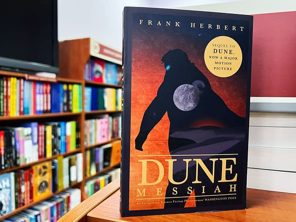A book cover for 'Dune Messiah' by Frank Herbert, featuring a silhouette of a figure against a red and orange background with celestial elements. The cover highlights its status as a sequel to 'Dune,' now a major motion picture, and is displayed in a colorful bookstore.