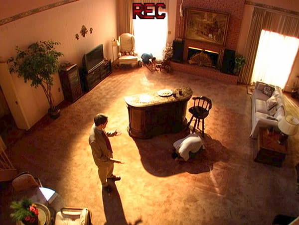 High-angle view of a warmly lit living room set with a fireplace, sofa, TV, and a central island, while two people appear to be cleaning or inspecting the floor. A REC graphic at the top suggests a recorded TV scene from Primetime.