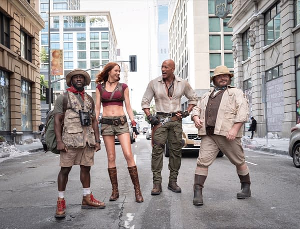 A scene from Jumanji 3 features four characters: Franklin 'Mouse' Finbar, Ruby Roundhouse, Dr. Smolder Bravestone, and Professor Shelly Oberon, standing in a city street, dressed in adventure-themed outfits ready for action.