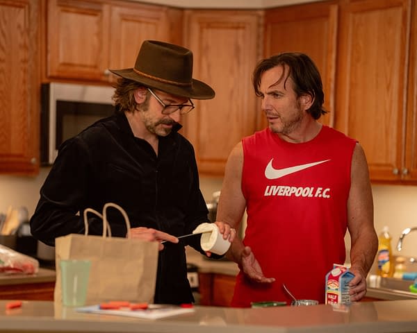 In a kitchen setting, two men are interacting; one wearing a black shirt and glasses holds a knife with a container, while the other in a red sleeveless shirt gestures with a milk carton. The scene captures a humorous moment from the film 'IS THIS THING ON?' featuring Bradley Cooper and Will Arnett.