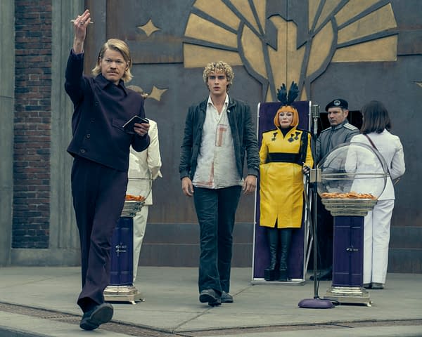 Jesse Plemons as Plutarch Heavensbee and Joseph Zada as Haymitch Abernathy stand before the Hunger Games reaping stage, flanked by officials in colorful Capitol costumes.